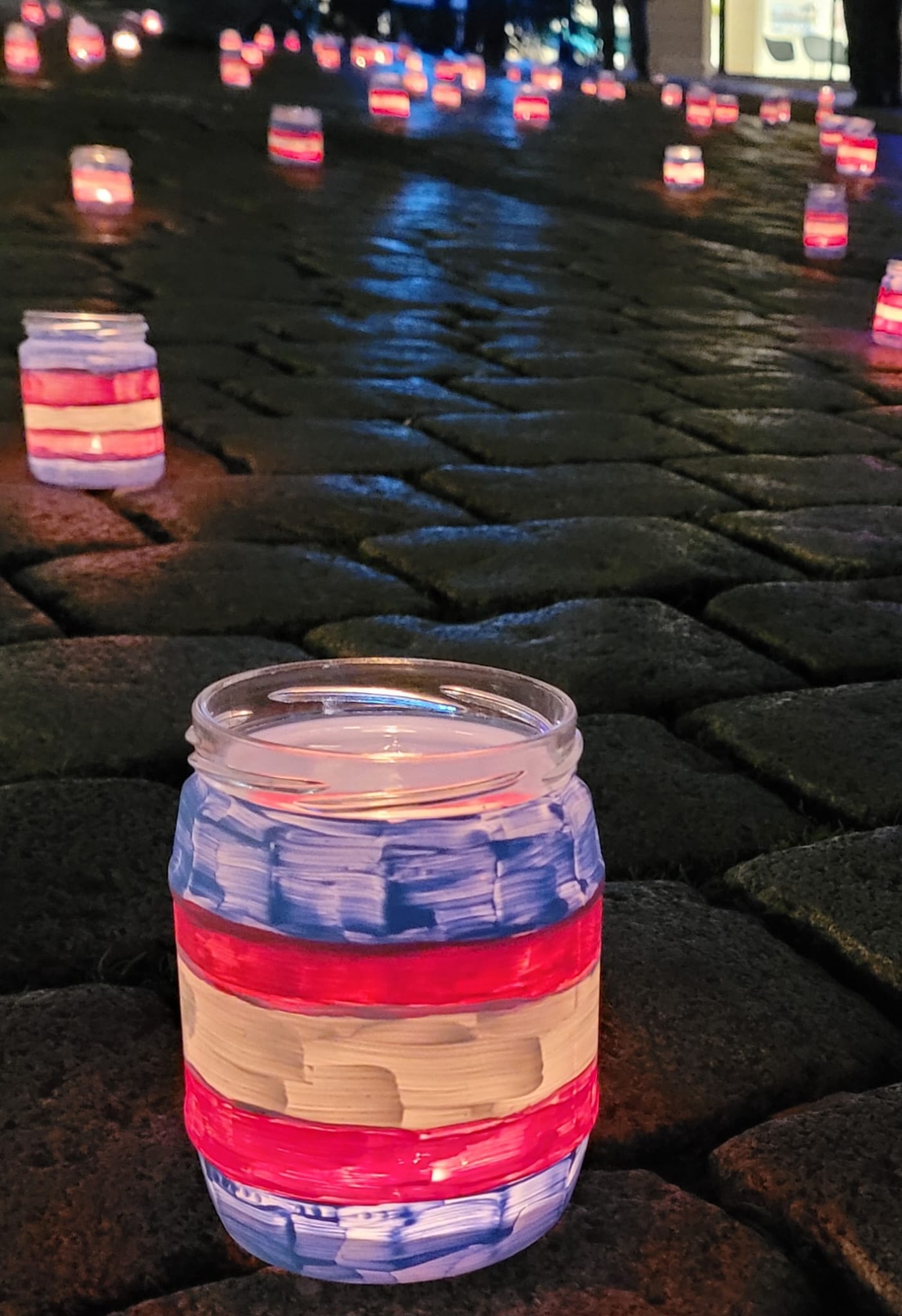 Teelichter in mit den trans Farben bemalten Gläsern bei Nacht auf Kopfsteinpflaster.